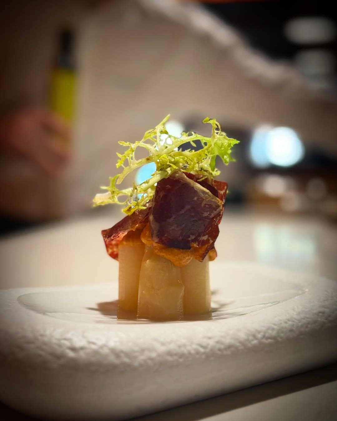 Espàrrec blanc gruixut tallat a trossos i dret sobre un plat blanc, coronat amb romesco, una làmina de cecina i escarola frisée.