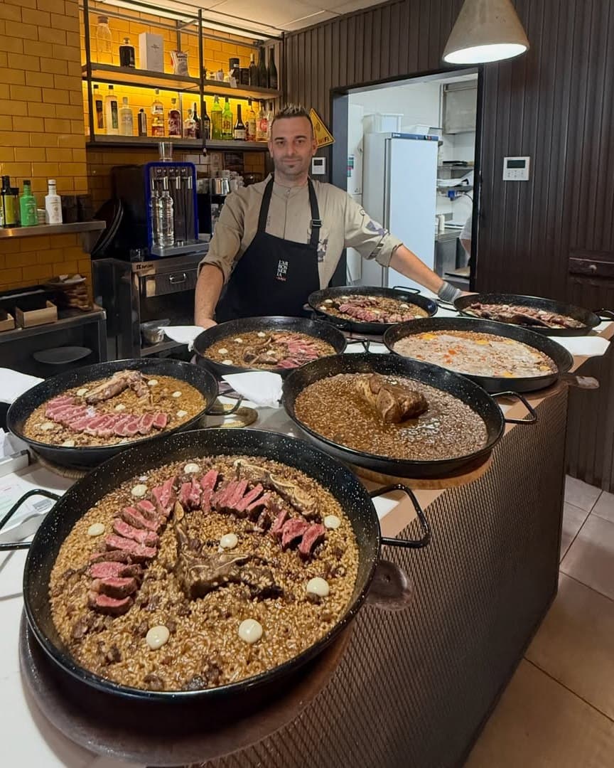 El xef Ramon Tudela, amb jaqueta beix i davantal negre, recolzat a la barra darrere de set paelles d'arròs acabades.